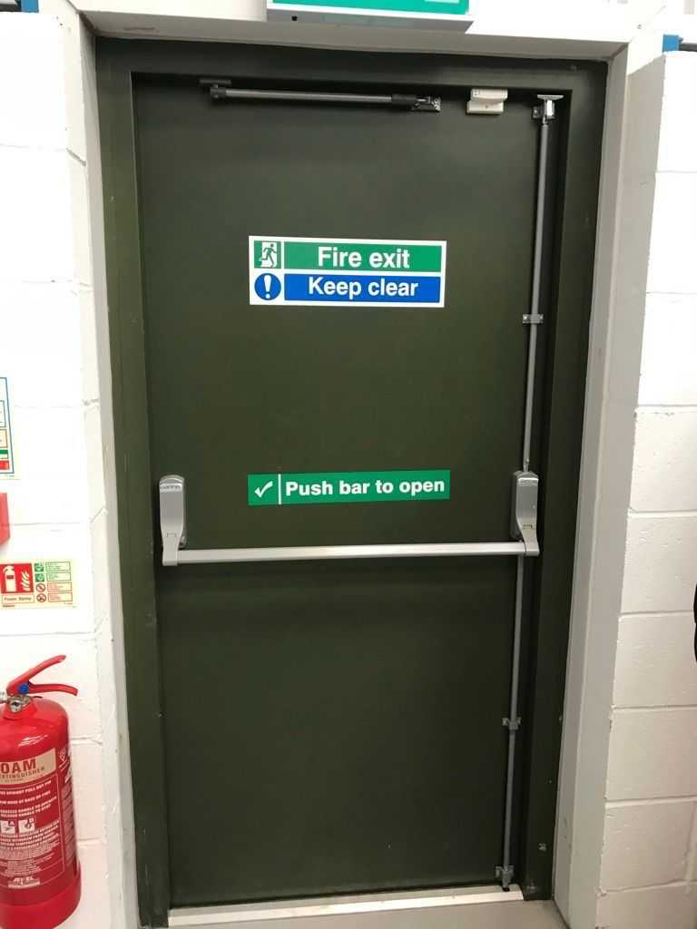 Green steel fire exit door with panic bar, vertical rods, door closer and fire exit signage in a commercial plant room