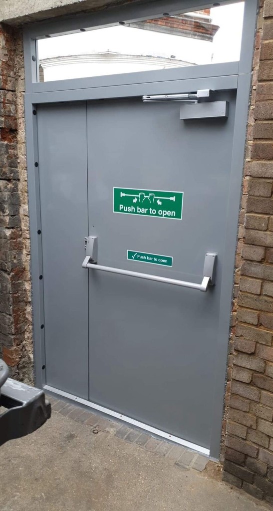 Single steel fire exit door with panic bar, glazed fanlight & fixed side panel at a commercial facility in North London