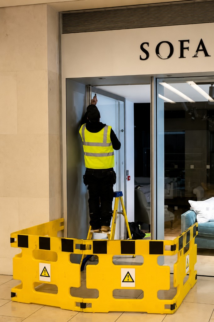 Engineer on a ladder repairing a commercial glass door closer behind a safety barrier at a retail entrance