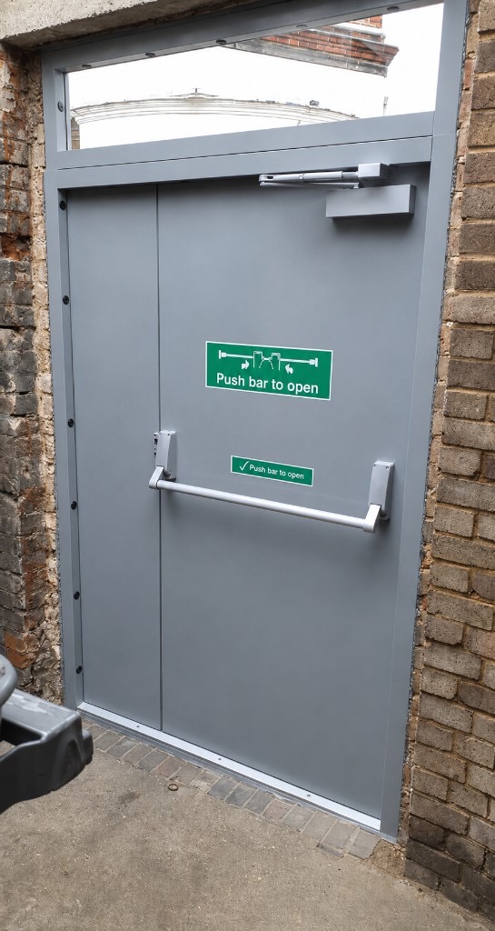 Overhead door closer mounted on a grey steel fire exit double door with panic bar
