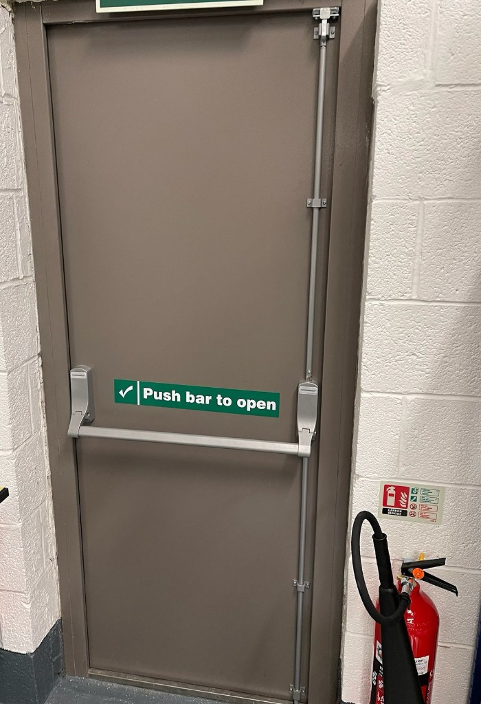 Brown steel fire exit door with push bar, vertical rods and emergency escape hardware in a commercial plant room