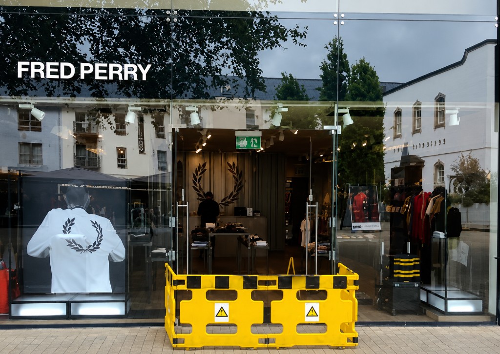 Frameless glass double doors with patch fittings and pull handles at a Fred Perry retail shopfront, with safety barrier during maintenance works